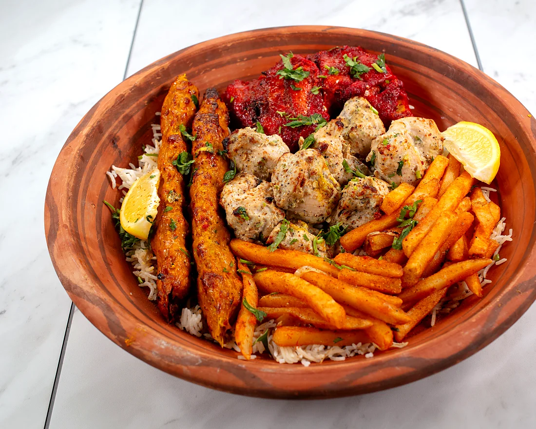 Clay bowl of mixed BBQ on rice: chicken seekh kababs, red chicken tikka, malai tikka, spiced fries and lemon wedges