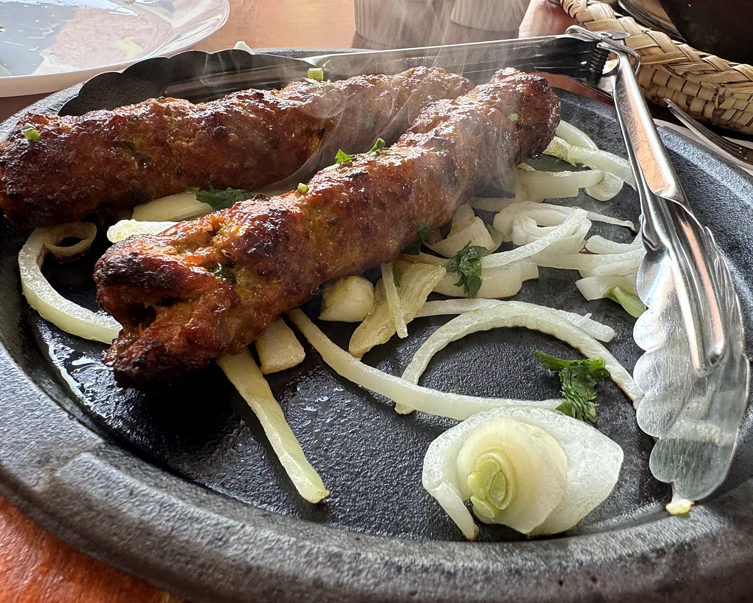 Two beef seekh kababs on a sizzling iron plate with onion rings and serving tongs
