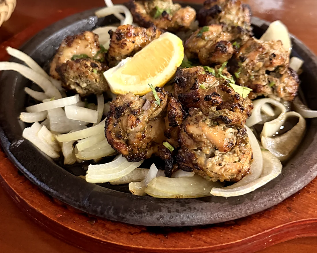 Grilled boneless chicken tikka pieces on a sizzling iron plate with onions and a lemon wedge