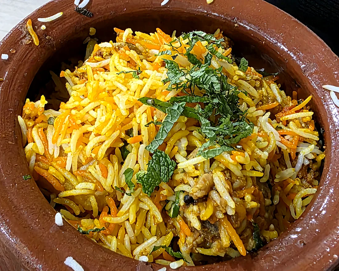 Biryani in a clay matka pot on a woven mat, saffron and white basmati rice topped with fresh mint
